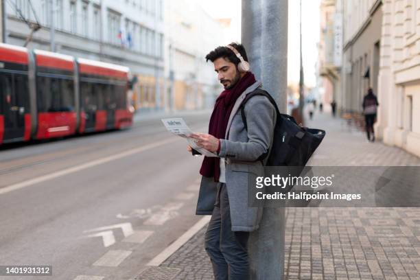 young handsome muslim man commuter waiting on the street, with backpack, headphones, reading newspaper. - man side way looking stock pictures, royalty-free photos & images