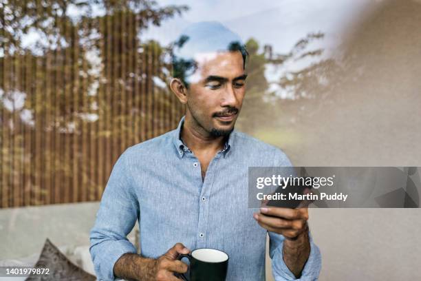 asian businessman working from home, using smartphone and drinking coffee, while standing at bedroom window of modern house - southeast asian people stock pictures, royalty-free photos & images