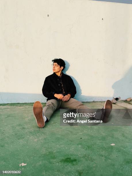 young man looking away while sitting with legs apart in front of wall - menswear stock-fotos und bilder