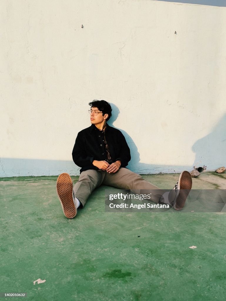 Young man looking away while sitting with legs apart in front of wall