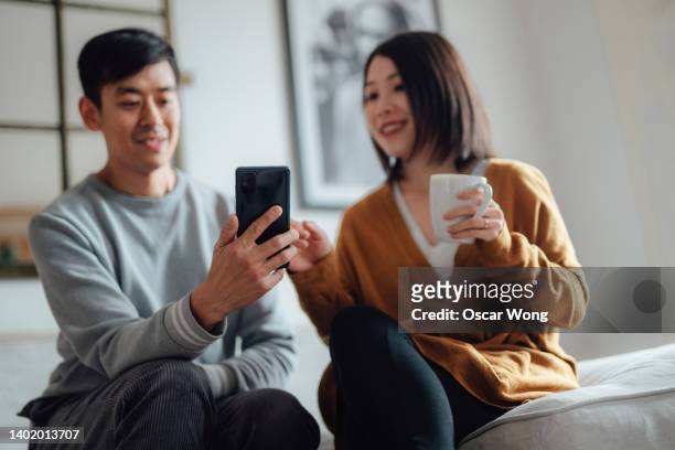 young asian couple shopping online with smart phone, sitting on sofa at home - asian married stock pictures, royalty-free photos & images