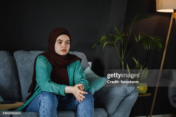 distraught young woman sitting on the sofa, hands clasped, contemplating - burka stock pictures, royalty-free photos & images