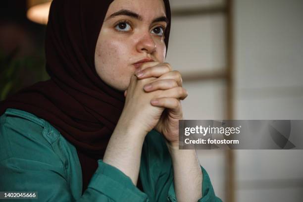 young woman in hijab sitting with hands clasped in front of face, contemplating - burka stock pictures, royalty-free photos & images
