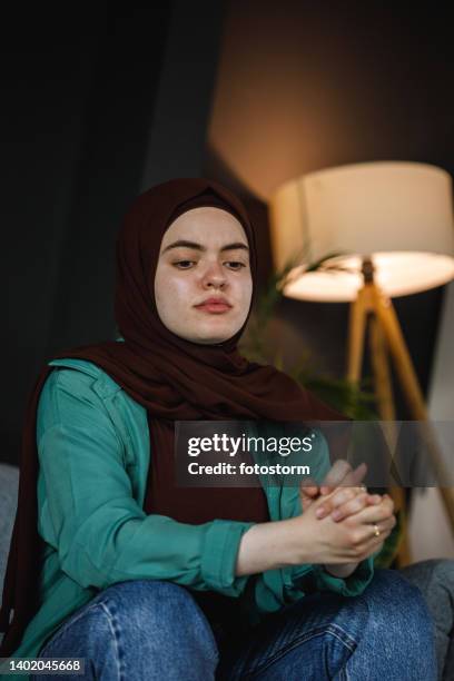 anxious young woman sitting with hands clasped, looking down, worrying, contemplating - burka stock pictures, royalty-free photos & images