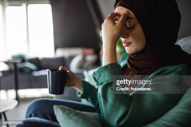 young woman rubbing her head due to headache while having a cup of coffee - burka stock pictures, royalty-free photos & images