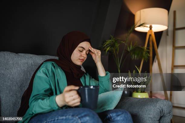 young woman rubbing her head due to a painful headache while relaxing, having a cup of coffee - burka stock pictures, royalty-free photos & images