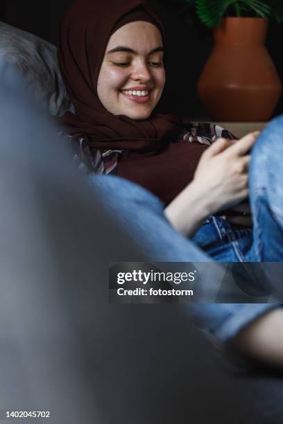 smiling young woman relaxing on the sofa and scrolling through memes online - burka stock pictures, royalty-free photos & images