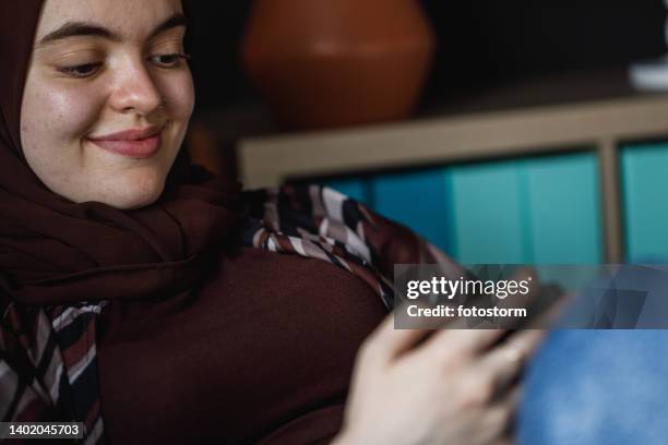 happy young woman lounging on the sofa and texting her crush via smart phone - burka stock pictures, royalty-free photos & images