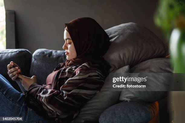 portrait of young woman lounging on the sofa and using smart phone - burka stock pictures, royalty-free photos & images