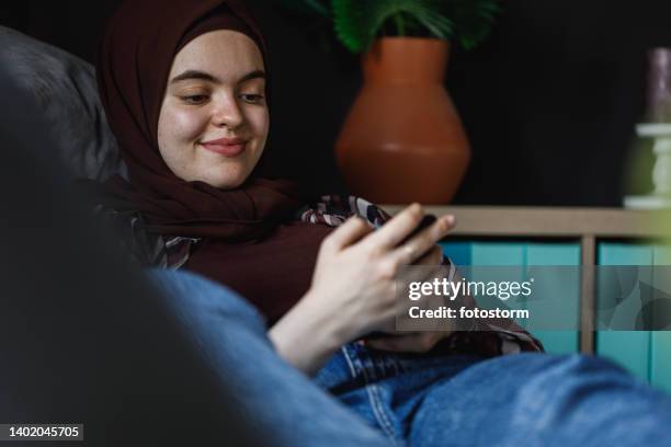 young woman lounging on the sofa and scrolling through memes her friends had sent her - burka stock pictures, royalty-free photos & images