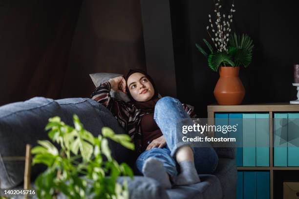 young woman lounging on the sofa in her living room, daydreaming - burka stock pictures, royalty-free photos & images