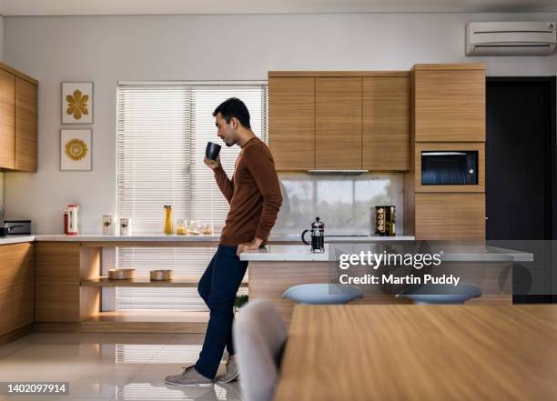 young asian man drinking coffee, leaning against modern kitchen island breakfast table with early morning sunlight - cozy kitchen stockfoto's en -beelden