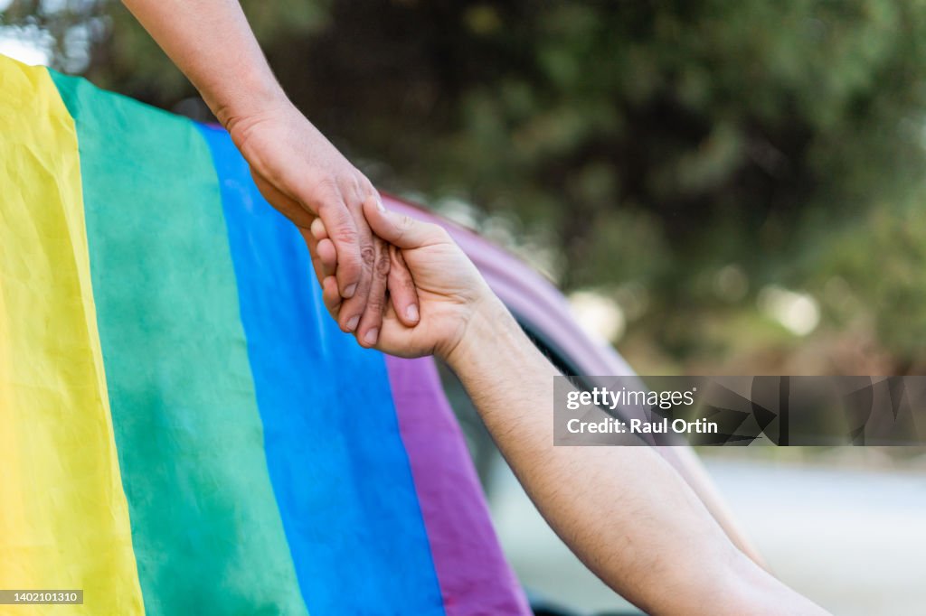 Unrecognizable gay couple holding hands with LGBT rainbow flag on background