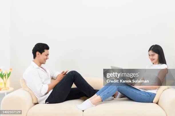 smiling young couple in living room at home using laptop and cell phone - couple seated apartment hipster imagens e fotografias de stock