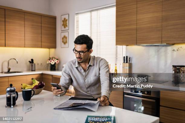 asian businessman  working from home, using smartphone and reading newspaper in modern kitchen at breakfast bar - wealthy asian man stock pictures, royalty-free photos & images