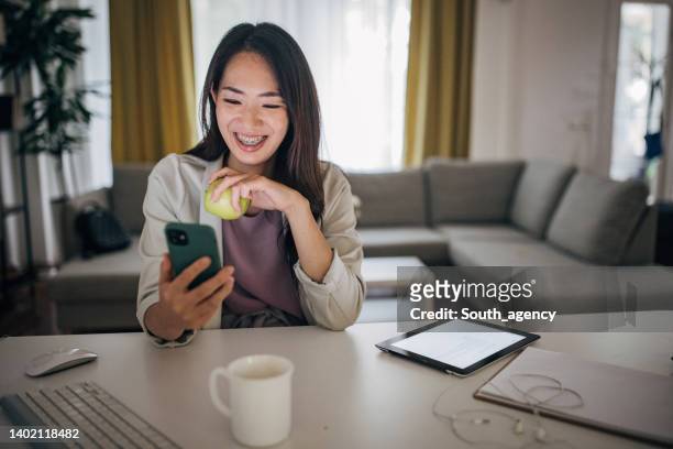 young woman using mobile phone while working at home office and eating apple - zuid europa stockfoto's en -beelden