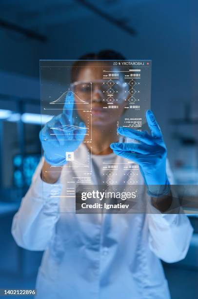 joven y hermosa médica afroamericana con bata de laboratorio y guantes protectores trabaja con una pantalla hud en un laboratorio futurista en una noche tardía - future health care fotografías e imágenes de stock