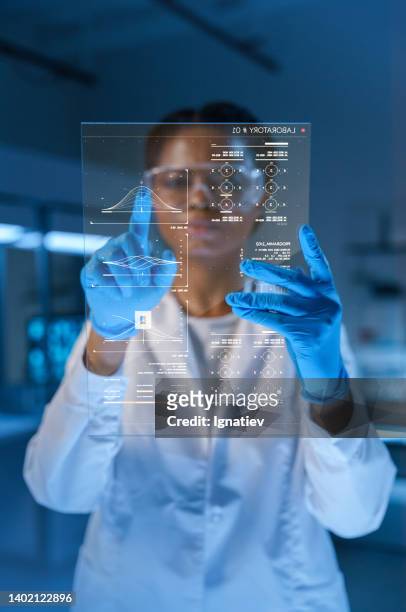 young beautiful african-american medic in a lab coat and protective gloves works with a hud screen in a futuristic lab in a late evening - future health care stockfoto's en -beelden