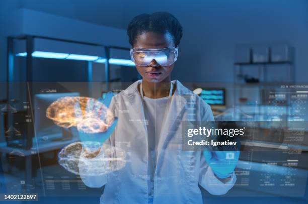 joven científica afroamericana con un atuendo médico, trabajando con una pantalla hud en un laboratorio futurista - anatomista fotografías e imágenes de stock