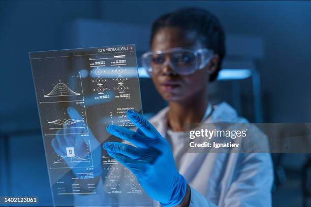 a young attractive female african- american doctor in a lab coat works with a small hud screen with diagrams in a modern laboratory - doctor lab coat bildbanksfoton och bilder