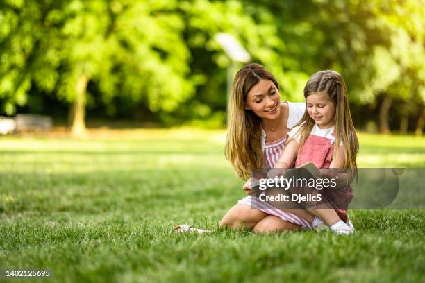 linda niña leyendo un libro con su madre en el parque. - mujer leyendo libro en el parque fotografías e imágenes de stock