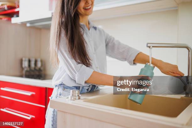 close up of smiling woman fills up reusable water bottle in the kitchen - filling stock pictures, royalty-free photos & images