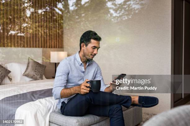young asian businessman working from home, using smartphone, sitting on bed at bedroom window of modern house - asian guy wake up stock pictures, royalty-free photos & images