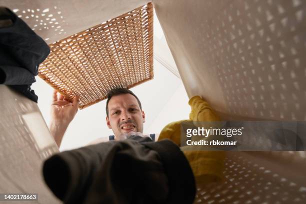 an unshaven man with a grimace on his face looks into a basket with dirty smelly laundry - onvolkomenheid stockfoto's en -beelden