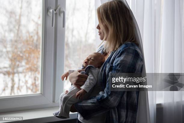 a young mother with her newborn baby in her arms stands by the window - cavan images stockfoto's en -beelden