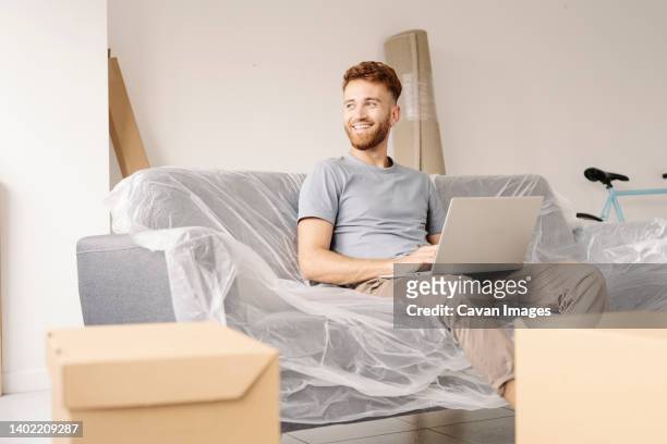 a young man sitting on his couch wrapped in plastic using his laptop. - man wrapped in plastic stock pictures, royalty-free photos & images