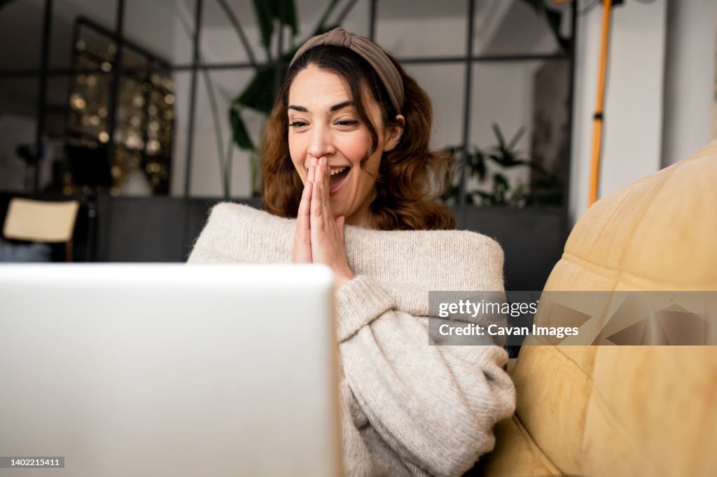 Excited woman looking at laptop screen sitting on cozy sofa at home