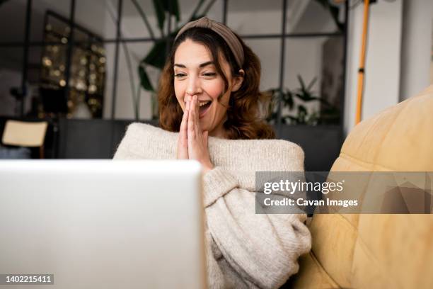 excited woman looking at laptop screen sitting on cozy sofa at home - joyeux photos et images de collection