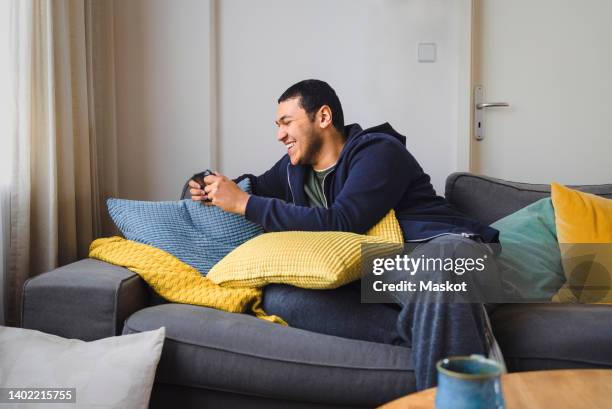 happy man playing handheld video game while sitting on sofa at home - gameboy stock-fotos und bilder