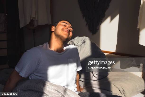 smiling man enjoying sunlight sitting on bed at home - aufstand stock-fotos und bilder