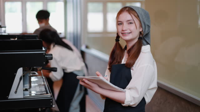 Young smiling student teenage girl learning to be barista while writing on notebook at coffee shop