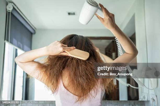 rear view of young asian woman using a comb for brushing her hair with a hair dryer for blowing water to dry her hair. - combing stockfoto's en -beelden