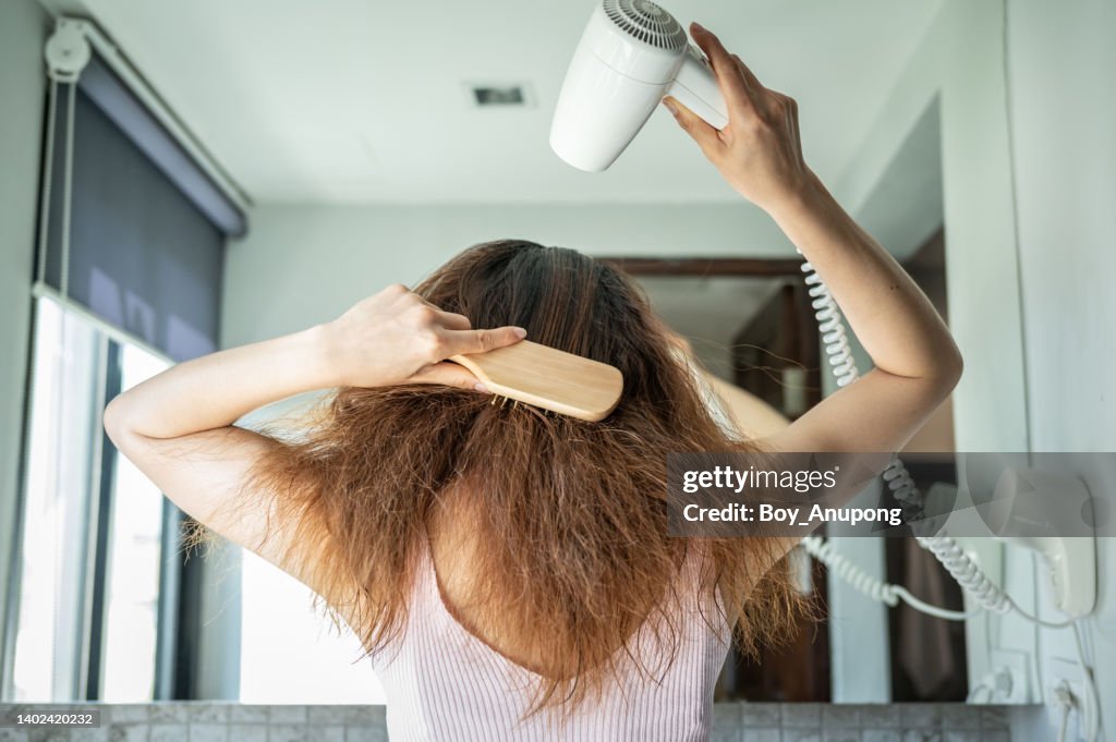 Rear view of young Asian woman using a comb for brushing her hair with a hair dryer for blowing water to dry her hair.