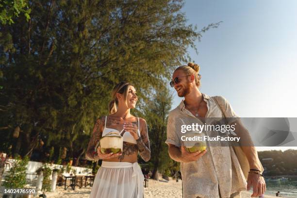 caucasian couple walking on the beach and drinking coconut on the beach - kokosmelk stockfoto's en -beelden