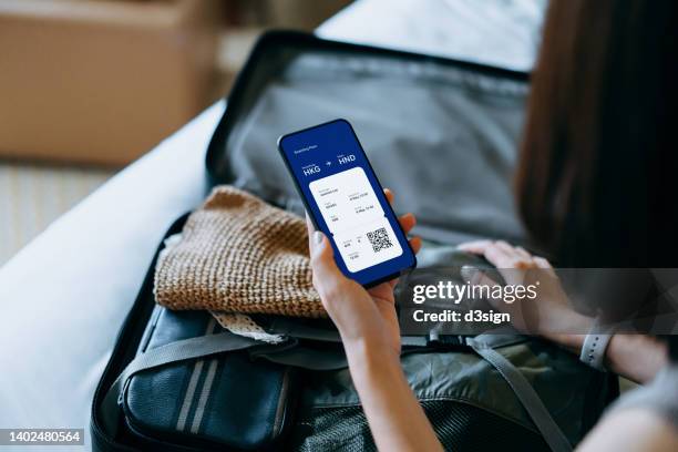 over the shoulder view of young asian woman using smartphone, checking digital flight ticket and boarding pass on device screen while packing a suitcase on bed for a trip. planning for travel. travel and vacation concept - booking hotel foto e immagini stock