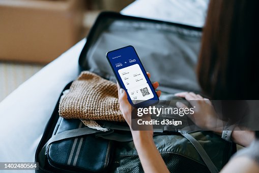 Over the shoulder view of young Asian woman using smartphone, checking digital flight ticket and boarding pass on device screen while packing a suitcase on bed for a trip. Planning for travel. Travel and vacation concept Over the shoulder view of young Asian woman using smartphone, checking digital flight ticket and boarding pass on device screen while packing a suitcase on bed for a trip. Planning for travel. Travel and vacation concept