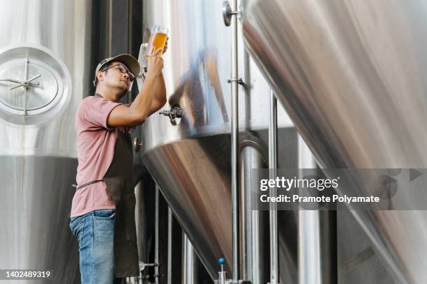 asian brewer filling beer in glass from tank at brewery in warehouse. family business concept. - bier brouwen stockfoto's en -beelden