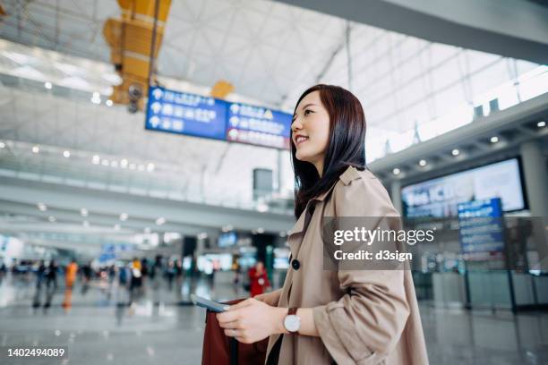 smiling young asian woman carrying suitcase and holding passport walking in airport terminal. ready to travel. travel and vacation concept. business person on business trip - pre reception stockfoto's en -beelden