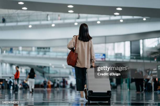 rear view of young asian woman carrying suitcase walking in airport terminal. ready to travel. travel and vacation concept. business person on business trip - travel insurance fotografías e imágenes de stock