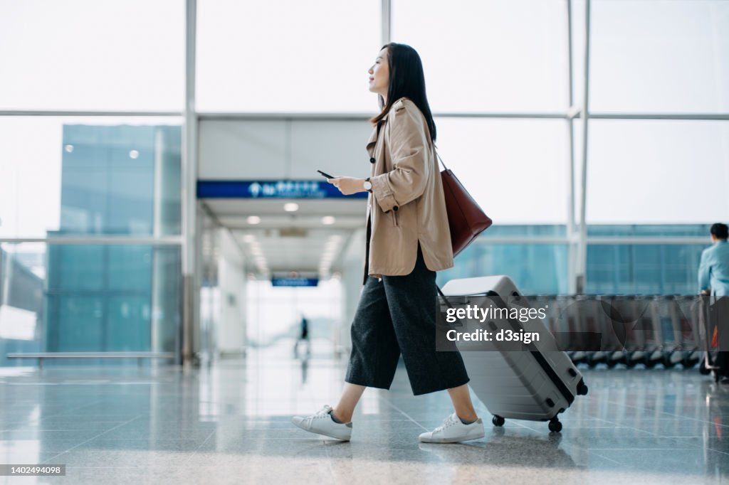 Young Asian woman carrying suitcase and holding smartphone on hand, walking in airport terminal. Ready to travel. Travel and vacation concept. Business person on business trip