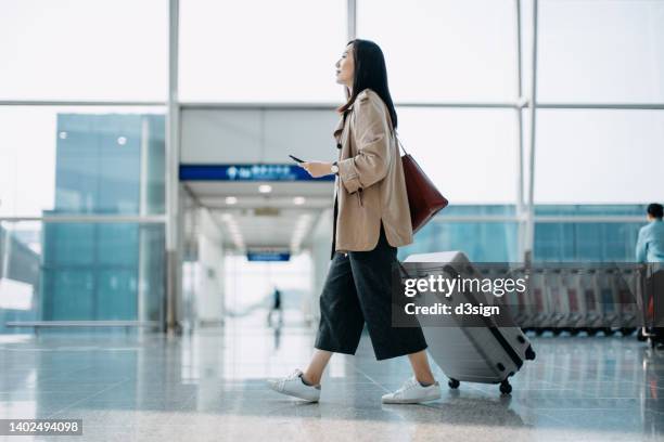 young asian woman carrying suitcase and holding smartphone on hand, walking in airport terminal. ready to travel. travel and vacation concept. business person on business trip - airport passenger foto e immagini stock