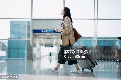 Young Asian woman carrying suitcase and holding smartphone on hand, walking in airport terminal. Ready to travel. Travel and vacation concept. Business person on business trip Young Asian woman carrying suitcase and holding smartphone on hand, walking in airport terminal. Ready to travel. Travel and vacation concept. Business person on business trip