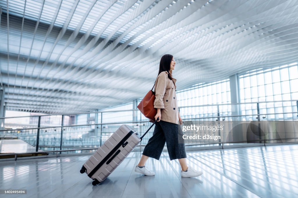 Side view of young Asian woman carrying suitcase walking in airport terminal. Ready to travel. Travel and vacation concept. Business person on business trip