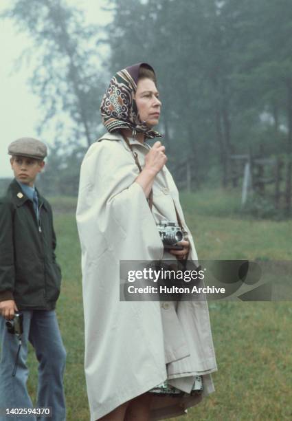 British Royals Edward, wearing a green waxed jacket and a flat cap, behind his mother, Queen Elizabeth II, in a beige raincoat and headscarf, a Leica...