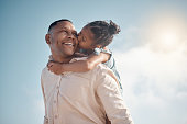 Smiling mixed race single father carrying little daughter on piggyback with copyspace. Adorable, happy, hispanic girl bonding with parent and kissing cheek on beach. Man and child enjoying free time