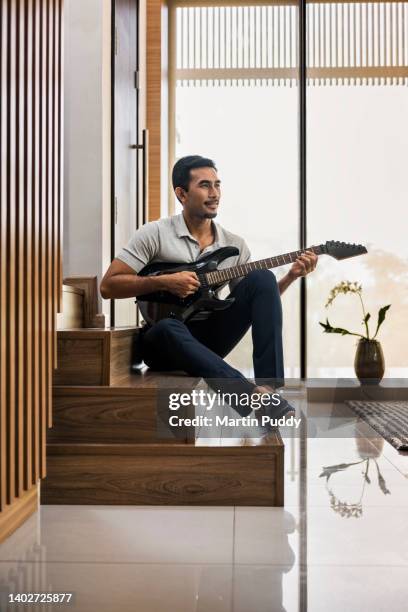 young asian man relaxing at home, playing guitar while sitting in modern living room - fabolous musician stockfoto's en -beelden